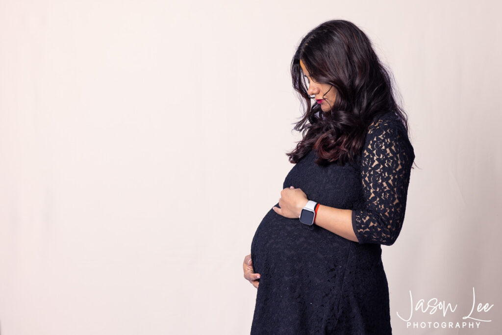 Woman in a black lace dress looking down while cradling her baby bump during a studio maternity session.