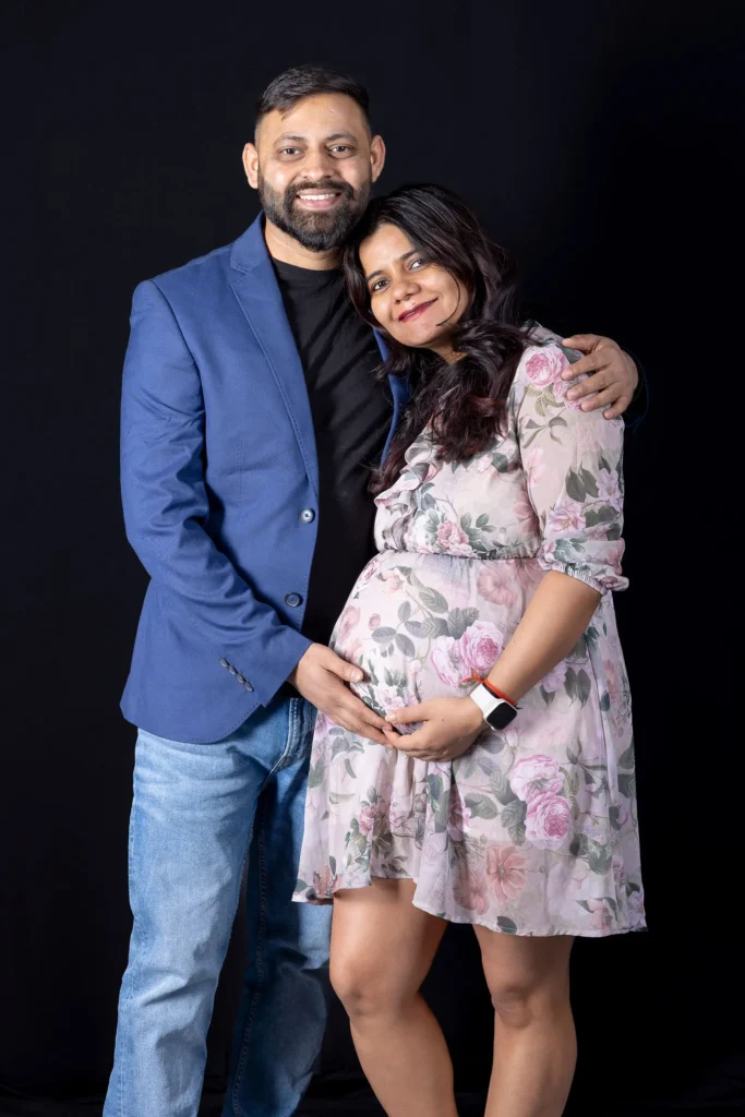Smiling couple with their hands together on the baby bump, wearing a floral dress and blue jacket.