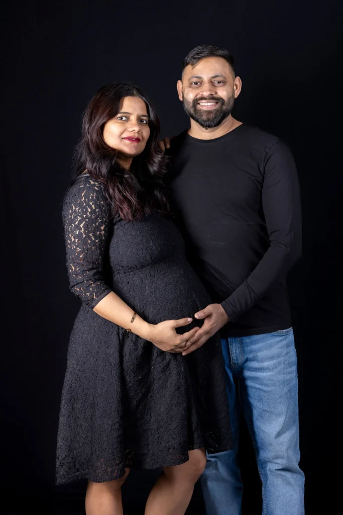 Expectant couple in black clothing with their hands on the baby bump against a dark backdrop.