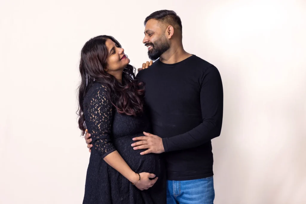 Couple smiling at each other with their hands on the baby bump against a pale backdrop.