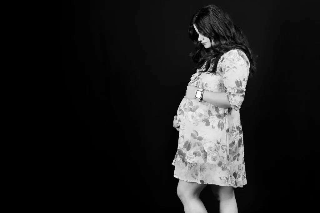 Black-and-white maternity portrait of a woman in a floral dress against a dark studio backdrop.