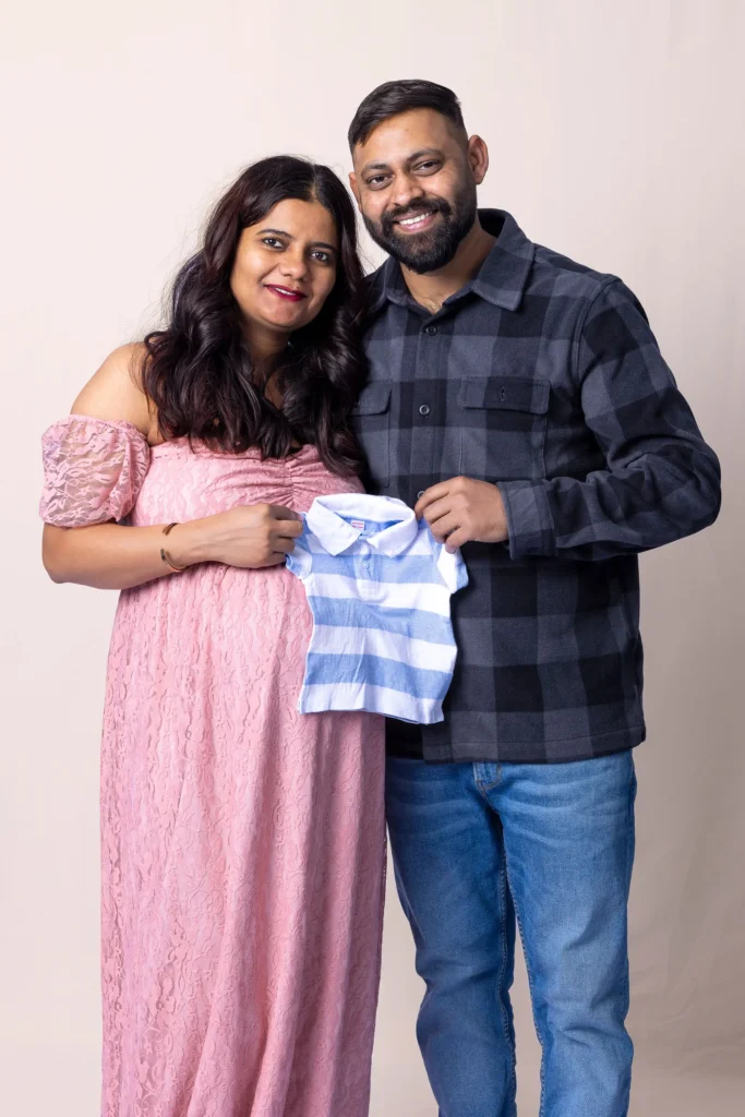 Smiling couple holding a tiny blue-and-white striped baby shirt during a maternity portrait.