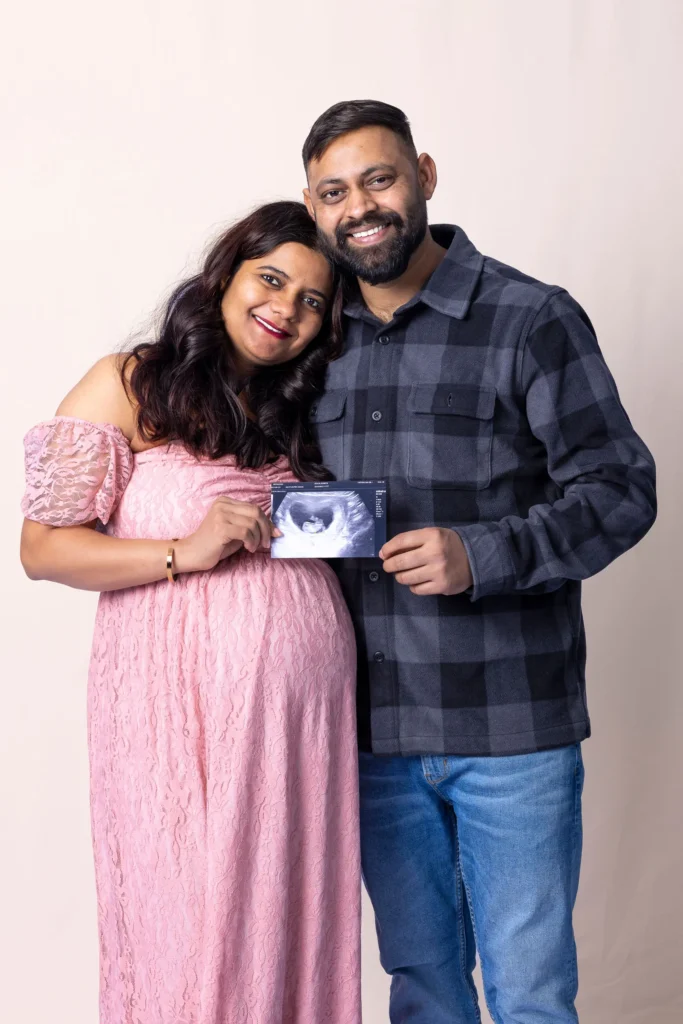 Smiling couple leaning together during a studio maternity portrait against a pale pink backdrop.