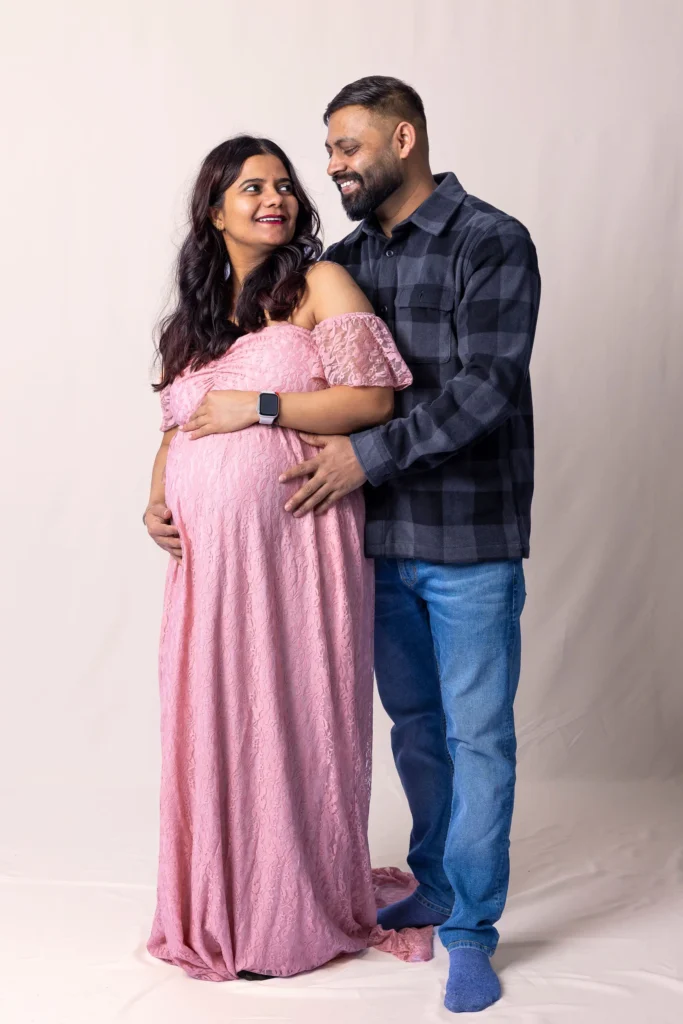 Man embracing his partner from behind as she rests her hands on her baby bump in a pink dress.