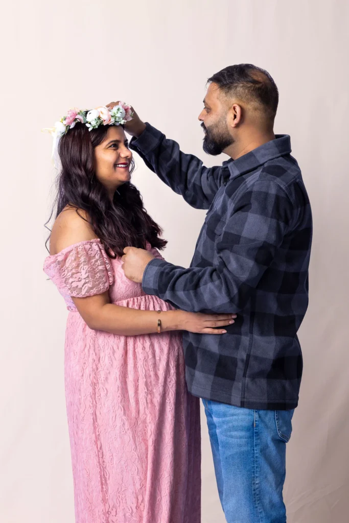 Man adjusting a flower crown on his smiling partner during a studio maternity session against a pale pink backdrop.