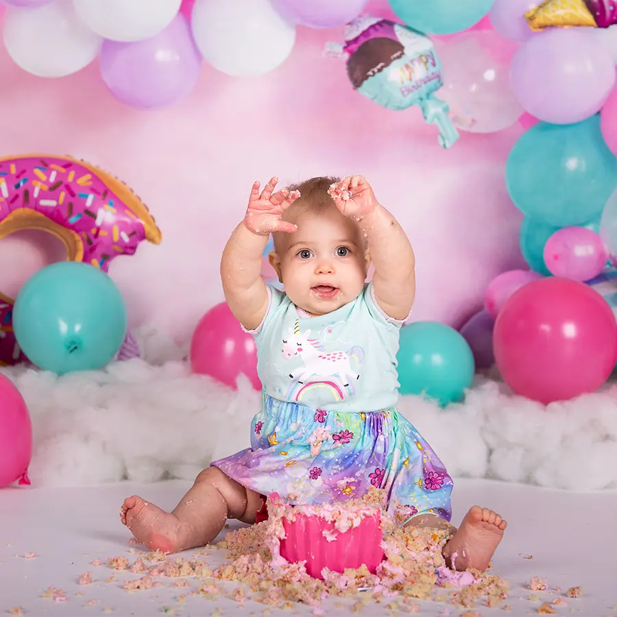 A one-year-old girl in a pink dress joyfully sitting with a giant pink cupcake smash cake in a bright, colorful studio backdrop in Winnipeg.