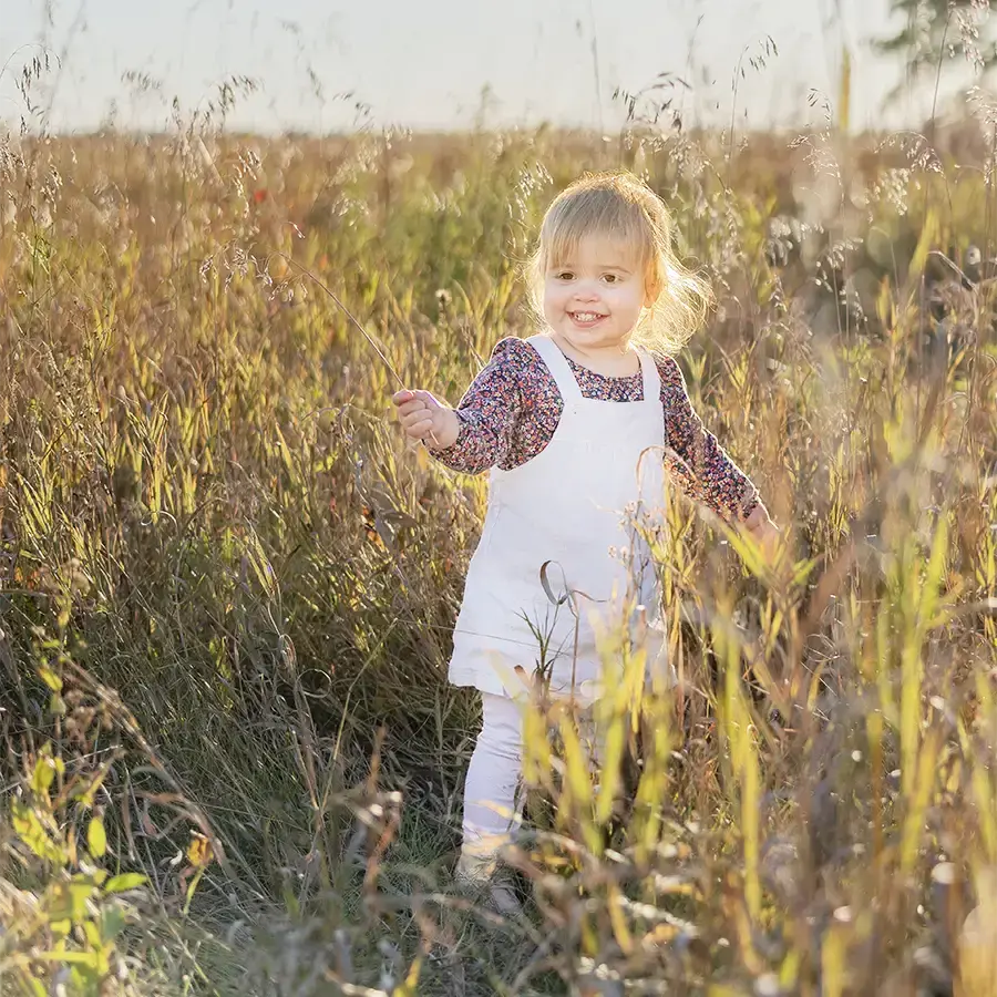 A toddler girl walking and smiling in the tall grass during golden hour at a Fall Family Session by Winnipeg Family Photographer Jason Lee