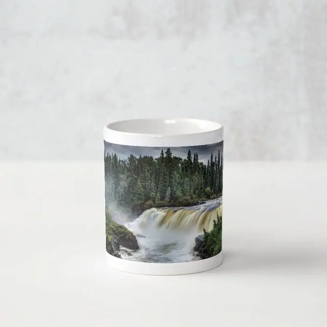 A white ceramic coffee mug with a panoramic photograph of Pisew Falls in Manitoba, by Métis photographer Jason Lee. The image shows rushing water flowing over granite cliffs.