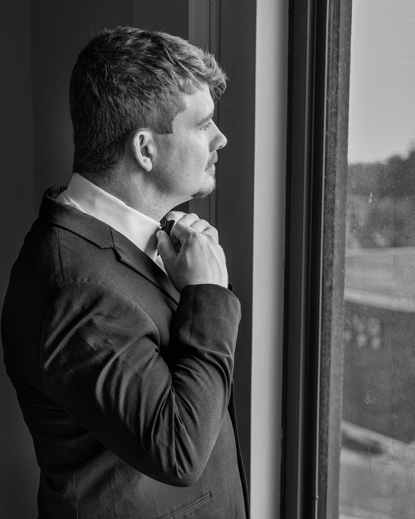 Groom adjusting his bow tie beside a window at the Viscount Gort Hotel