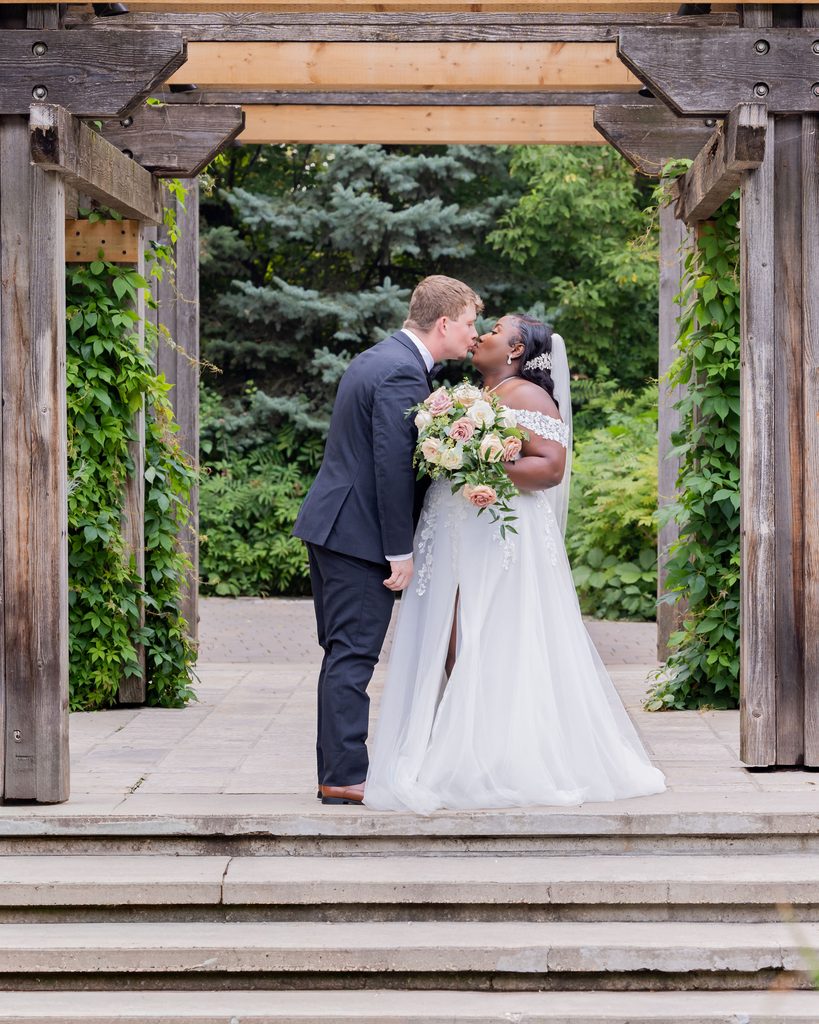 Newlyweds kissing beneath the pergola at Leo Mol Sculpture Garden