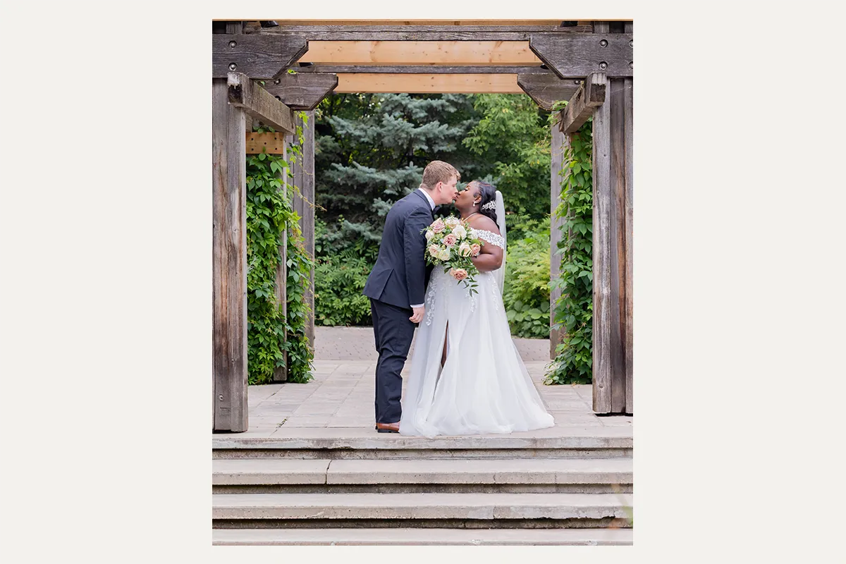 Bride and groom kissing beneath a wooden garden pergola during their wedding portraits