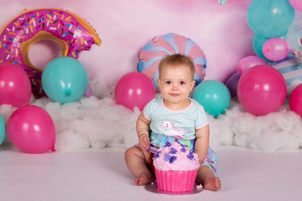 Baby sitting behind a pink cupcake cake on a colourful birthday set