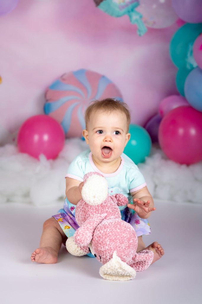 Baby sitting with a pink stuffed animal before a first birthday cake smash