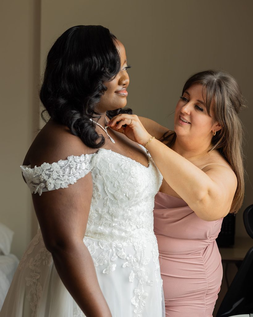 Maid of honour helping the smiling bride adjust her necklace before the wedding.