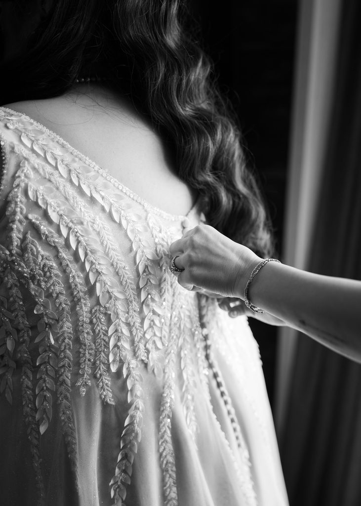 Black and white close-up of a hand fastening the bride’s lace wedding dress before her ceremony at the Victoria Inn.