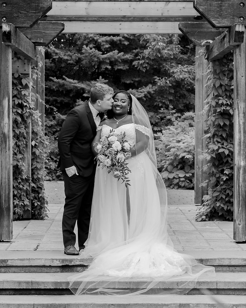 Black and white portrait of the groom kissing the bride’s cheek beneath a pergola in Assiniboine Park.