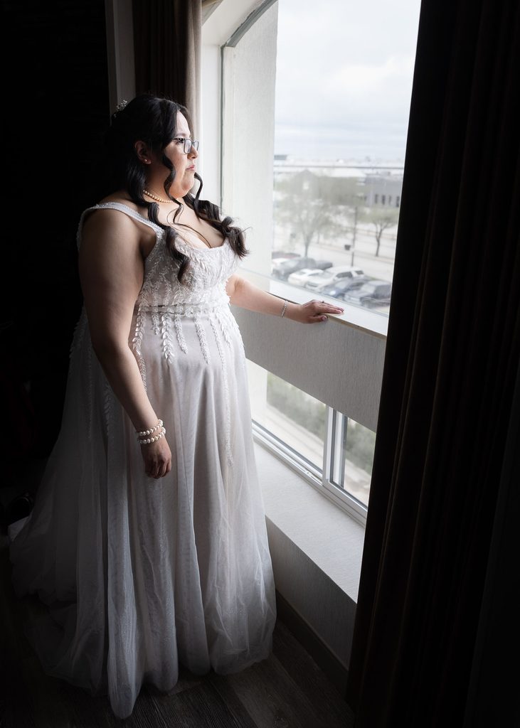 Bride in a lace wedding dress looking out of a bright window before the ceremony.