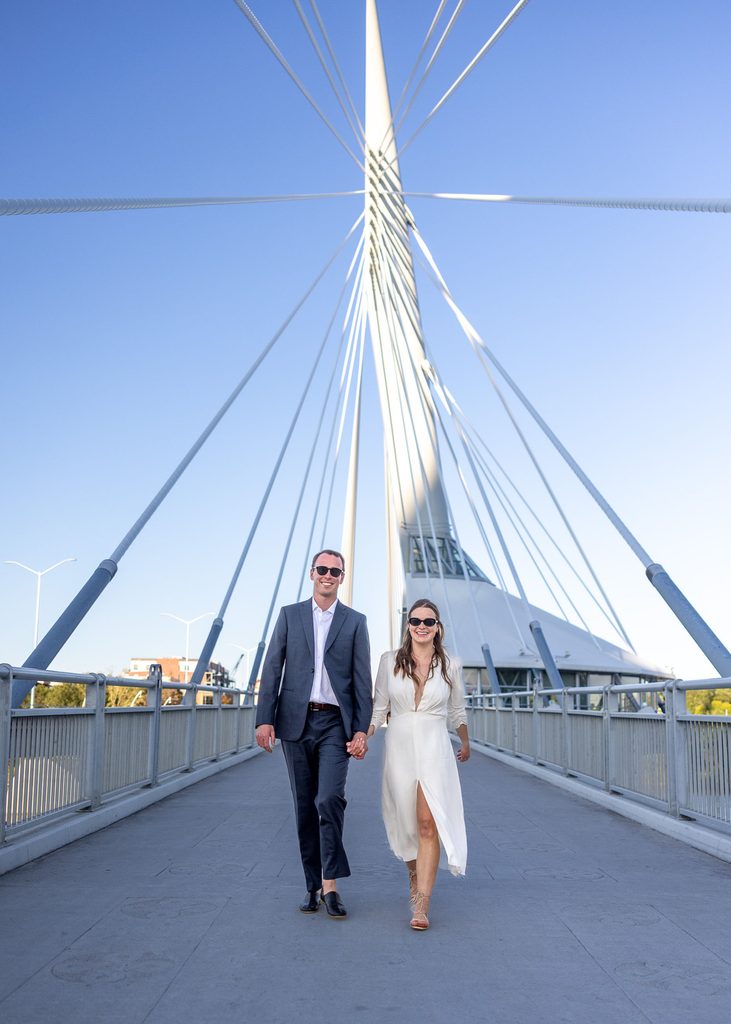 Bride and groom wearing sunglasses and walking hand in hand on the Esplanade Riel bridge in Winnipeg.