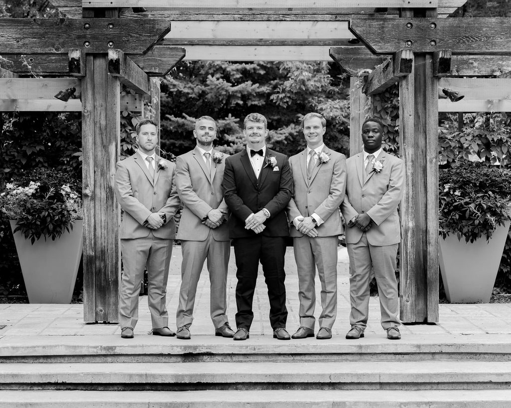 Black and white portrait of the groom and four groomsmen standing beneath a wooden pergola.