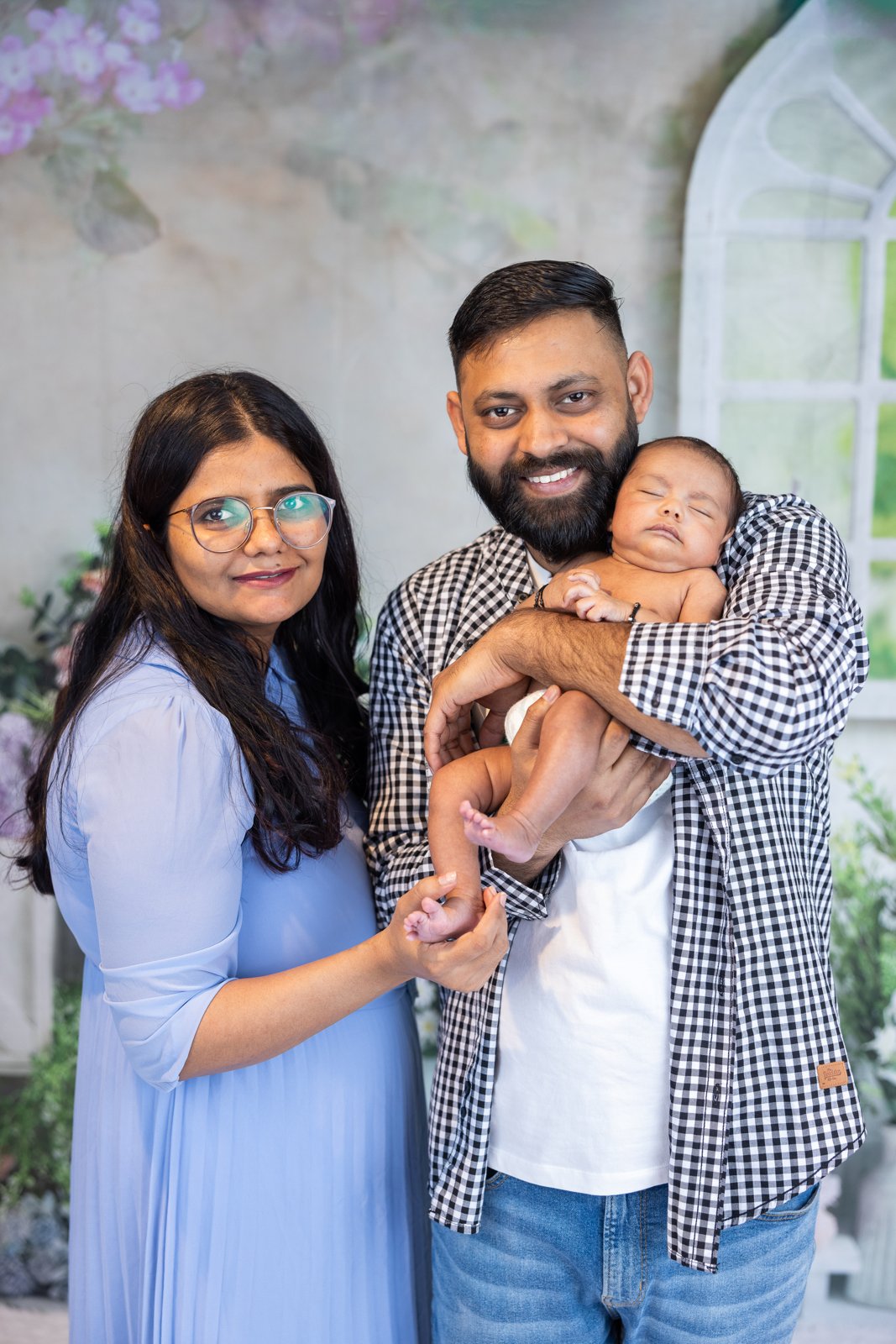 Parents standing together with their newborn against a floral studio backdrop.