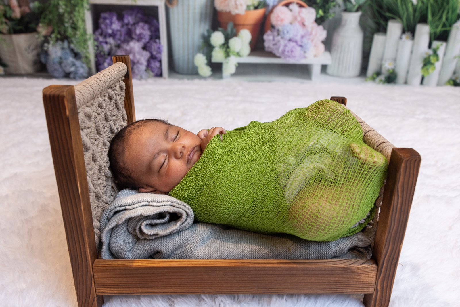 Newborn in a green wrap resting in a small wooden bed prop.