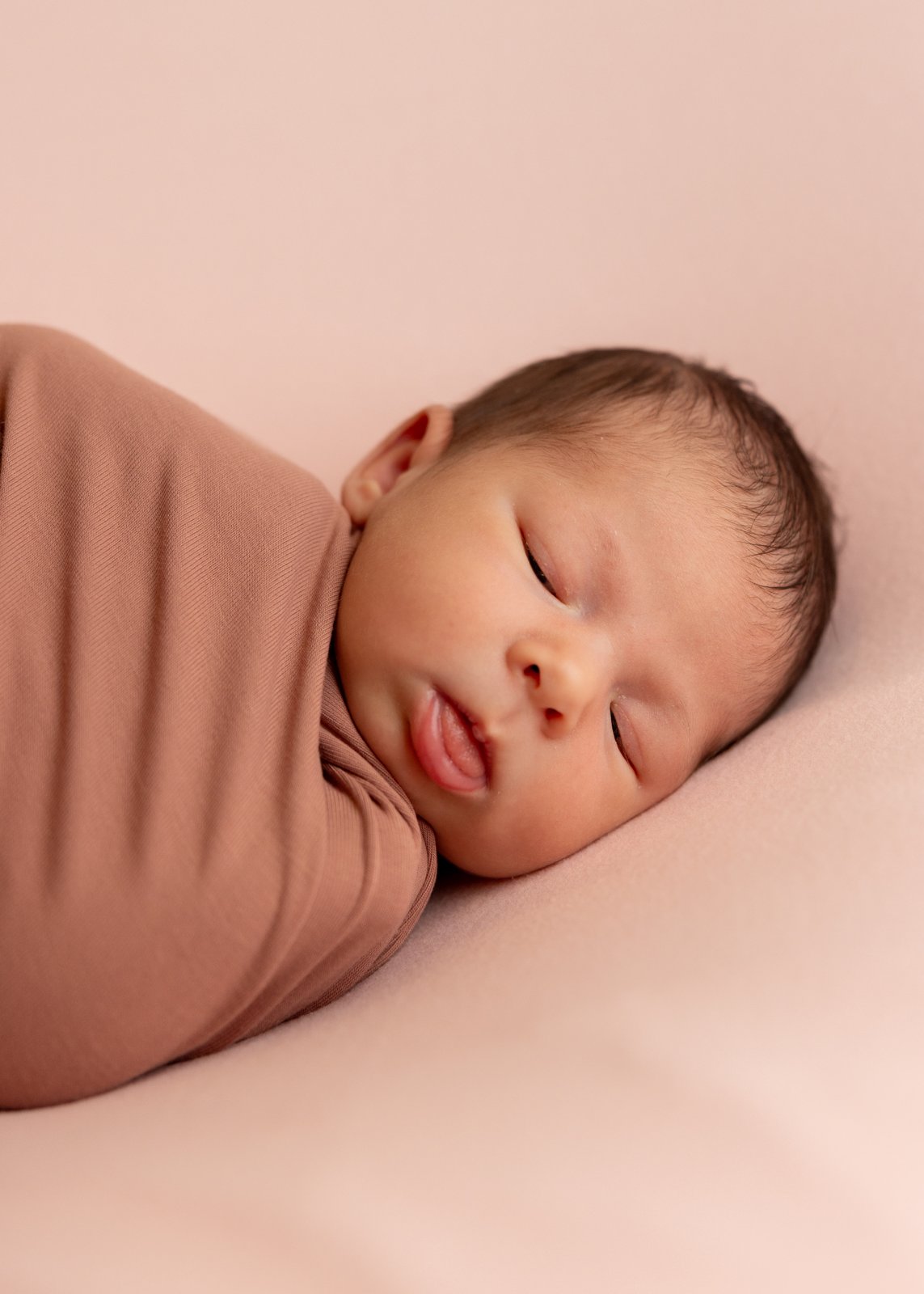 Sleeping newborn wrapped in warm pink fabric on a pale pink blanket.
