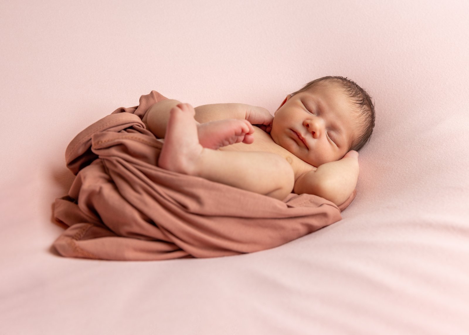 Sleeping newborn with one hand beside their face on a pink blanket.